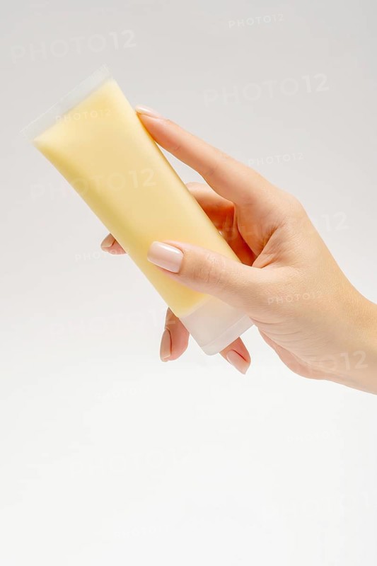 A close-up view showcasing of a cropped unrecognizable hand holding a tube of yellow facial mask, designed for anti-aging care with ingredients like manuka honey. The background is plain, highlighting the product