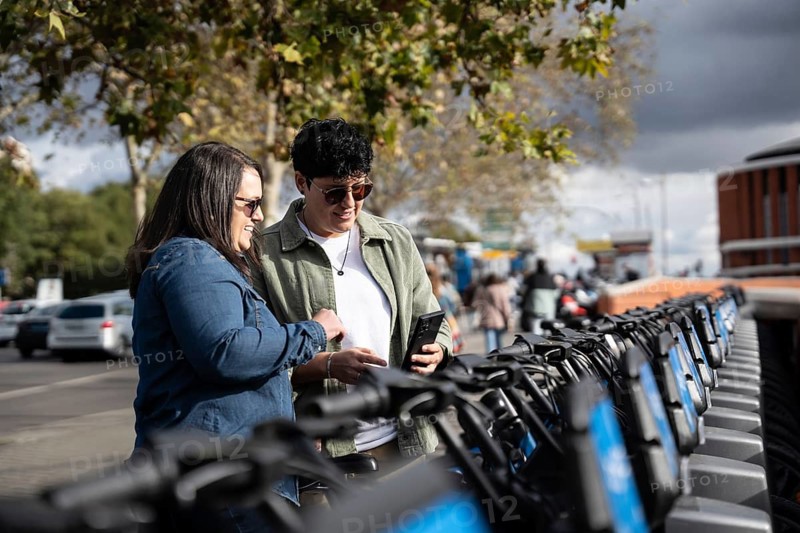 A joyful lesbian couple explores a row of rental bikes in a vibrant city setting. Background features a charming mix of city life and nature, highlighting urban exploration
