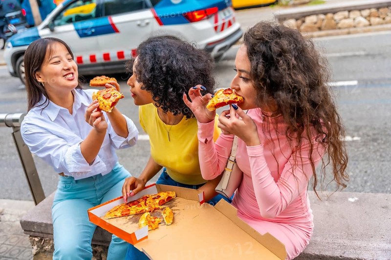 Three multiracial women sharing pizza sitting together in the street