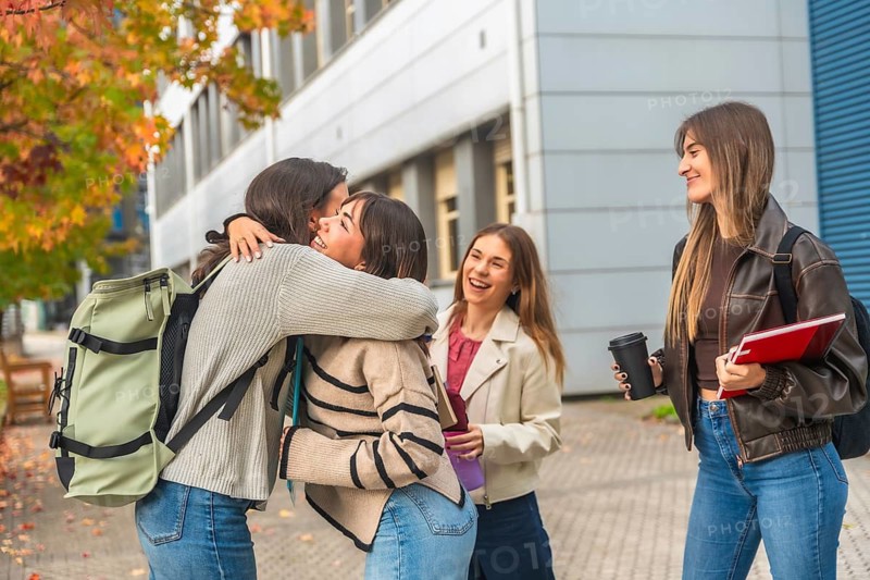 Four cheerful young women students hug and laugh on a university campus amid autumn leaves, reuniting between classes with backpacks, books and warm smiles