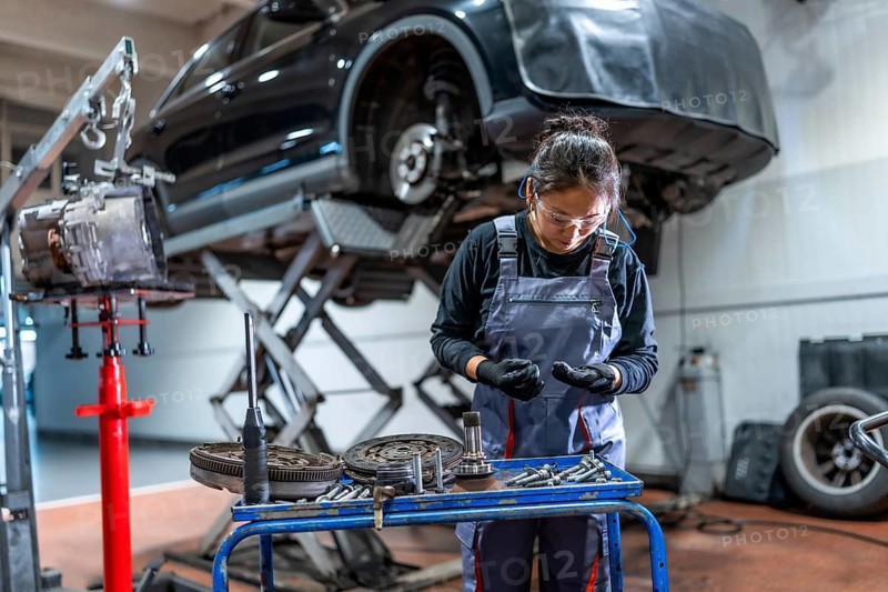 Female mechanic wearing gloves and safety glasses examining car parts with a car on a hydraulic lift in the background