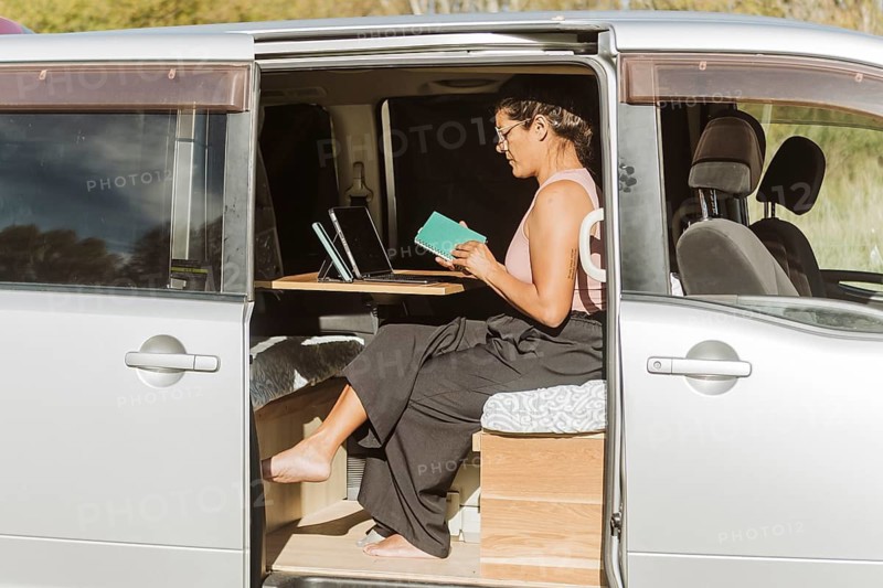A woman works inside a van, utilizing a laptop and notebook to stay productive amidst the vibrant autumn scenery of New Zealand. The open van door reveals a cozy workspace