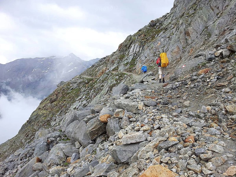Two hikers with large backpacks climb a rocky mountain trail surrounded by fog, E5, hiking in the Alps, crossing the Alps, Austria