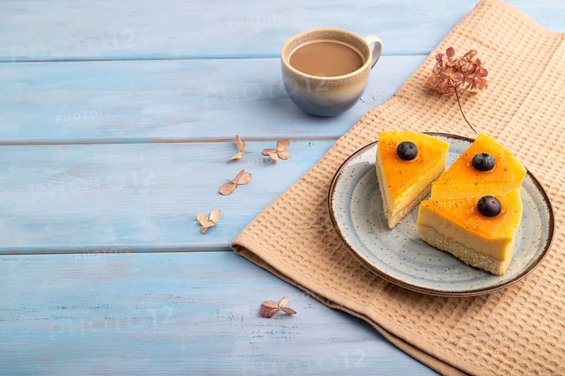 Cheesecake with mango cream, cup of coffee, on blue wooden background with beige textile, copy space, side view