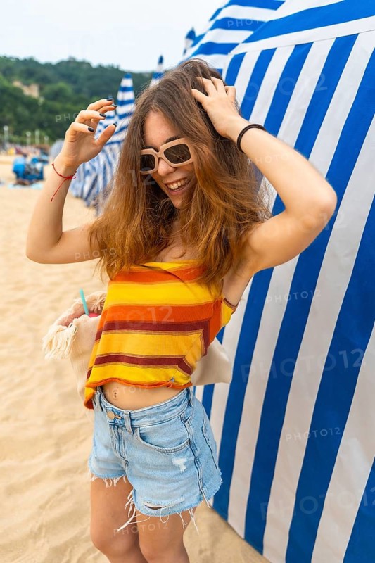 Young woman smiling with sunglasses, wearing a striped top and denim shorts, standing on a sandy beach next to blue and white striped cabanas, enjoying summer vacation