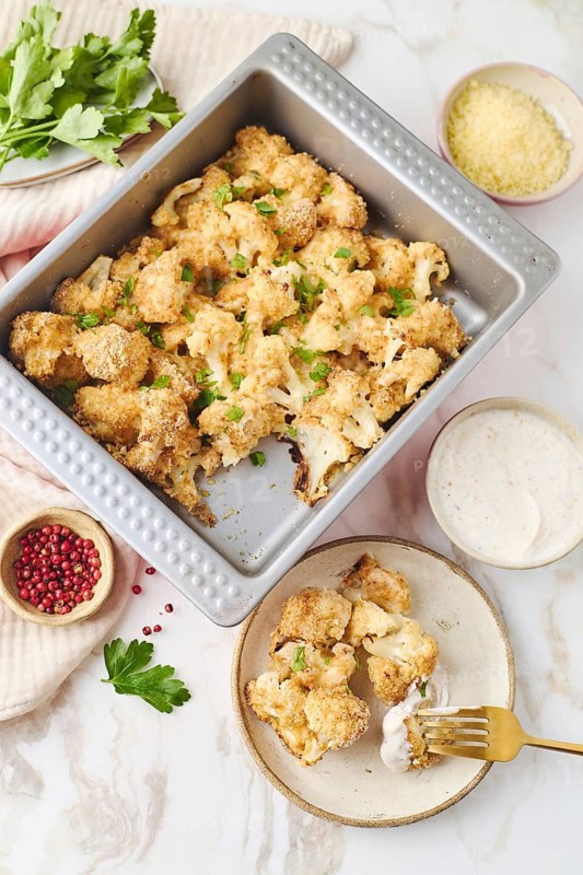 Top view of savory baked cauliflower in breadcrumbs, coated with egg and sprinkled with parsley and cheese, served with cream sauce in a metal baking form