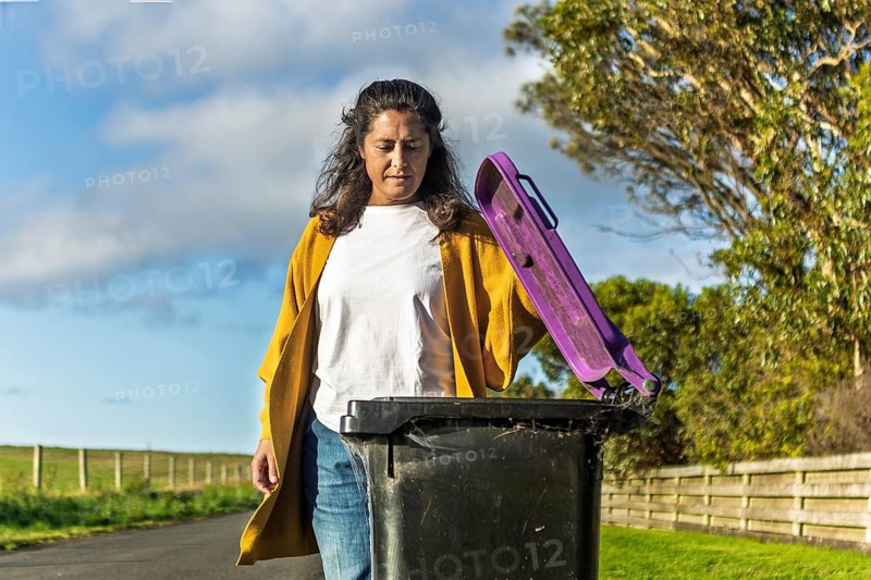A woman in casual attire handles a bin outside her Australian home. The scenic background highlights a typical day in suburban life, focused on responsible living