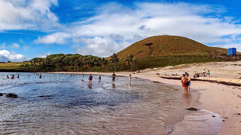 People playing and relaxing on a beach lined with palm trees and hills, the landscape of Easter Island in the Pacific Ocean