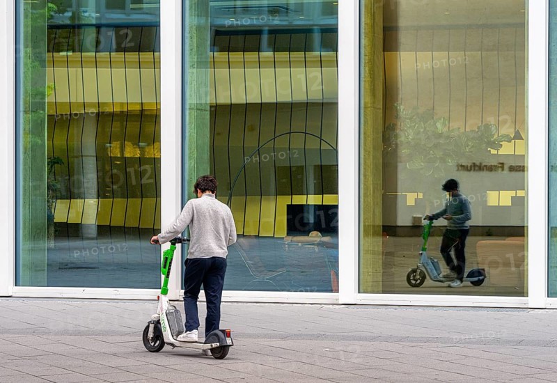 Young man with an electric scooter standing in front of the entrance to the One office building in downtown Frankfurt, Hesse, Germany