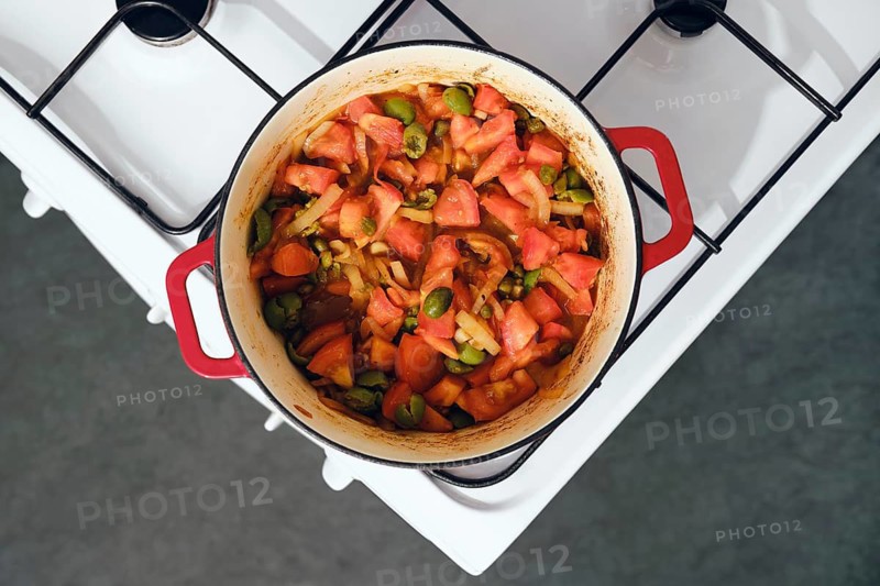 Red cast iron pot sits on white stove. Inside pot are chopped tomatoes, onion, capers and green olives, all mixed together. This scene takes place in kitchen during daytime