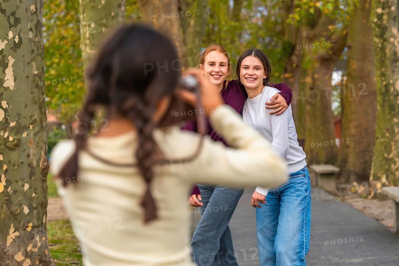 Young girl holding a camera. Capturing a moment with two smiling teenage sisters embracing each other outdoors in an autumn park setting. Representing family connections. Friendship. And leisure time