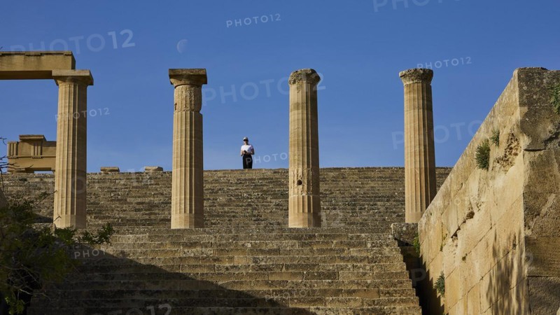 A person stands between ancient columns under a blue sky with visible moon, Acropolis of Lindos, morning light, Propylaea with staircase, Lindos, Rhodes, Dodecanese, Greek Islands, Greece
