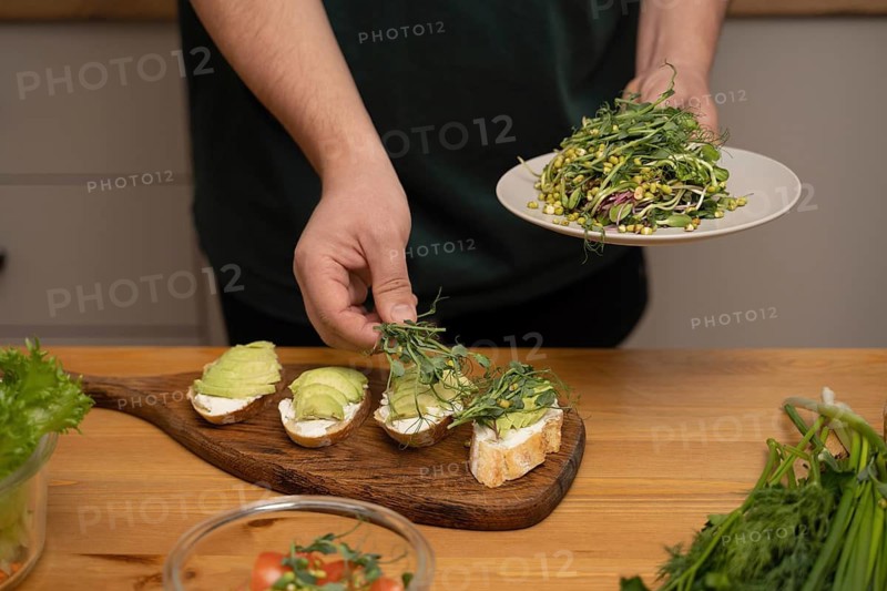 A person prepares avocado toast topped with cream cheese and fresh greens on a wooden board. A kitchen scene showcasing delicious and healthy homemade snacks