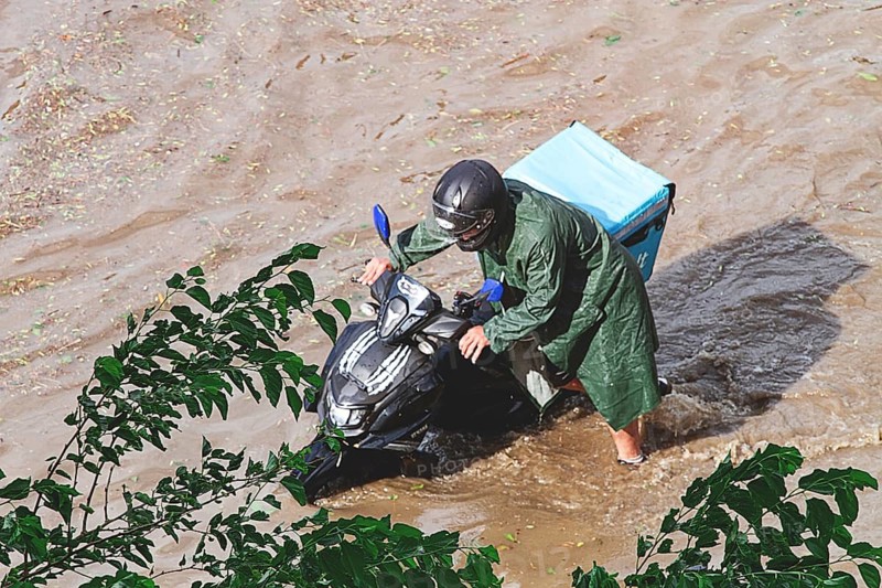 A delivery person is walking next to a motorcycle that is half-submerged in water. The street is flooded due to heavy rain. People may notice the challenging conditions for deliveries