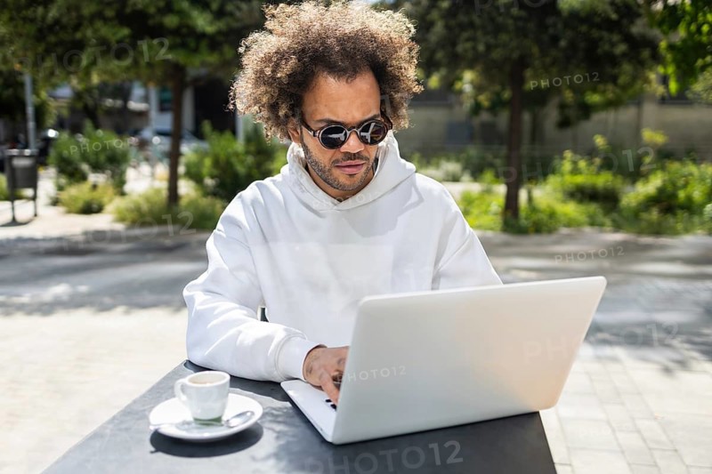 A businessman with afro hairstyle, wearing sunglasses and a white sweatshirt, works on a laptop at an outdoor cafe. A cup of coffee sits nearby, with trees in the background