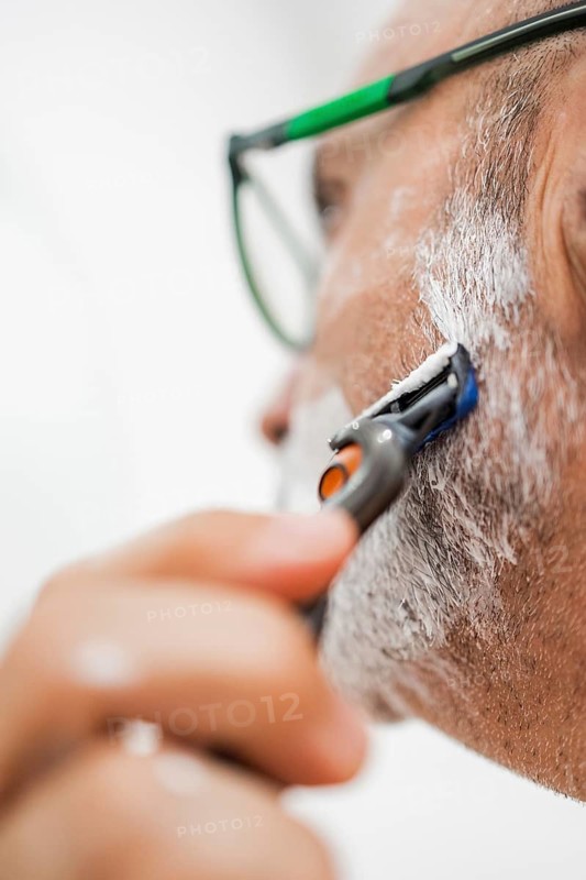 Close-up of a mature man shaving in the bathroom. The man, wearing glasses, uses a razor across a lathered face, showcasing a daily grooming routine