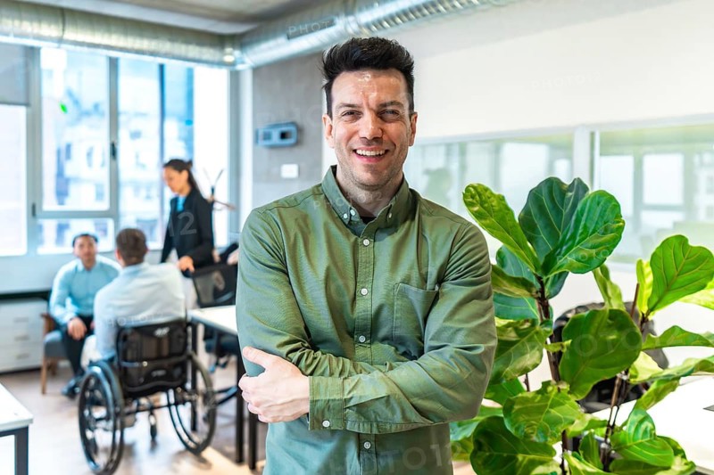 Smiling businessman with crossed arms stands in a modern office, collaborating with colleagues, including a person in a wheelchair
