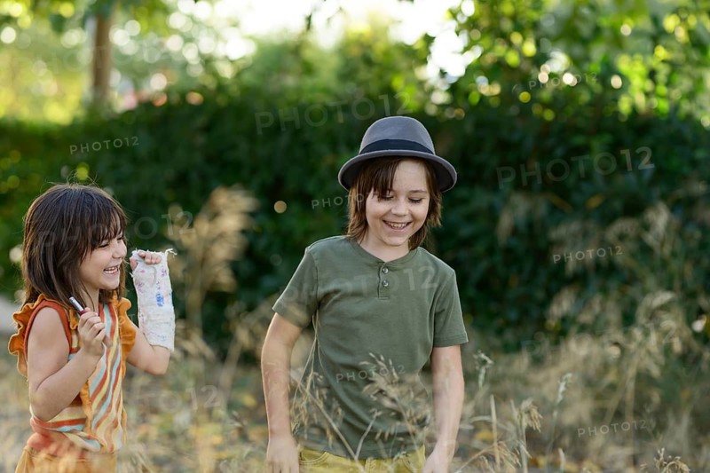 Two children joyfully explore a garden, surrounded by lush greenery. One wears a hat, while the other displays a creative cast on her arm, showcasing creativity