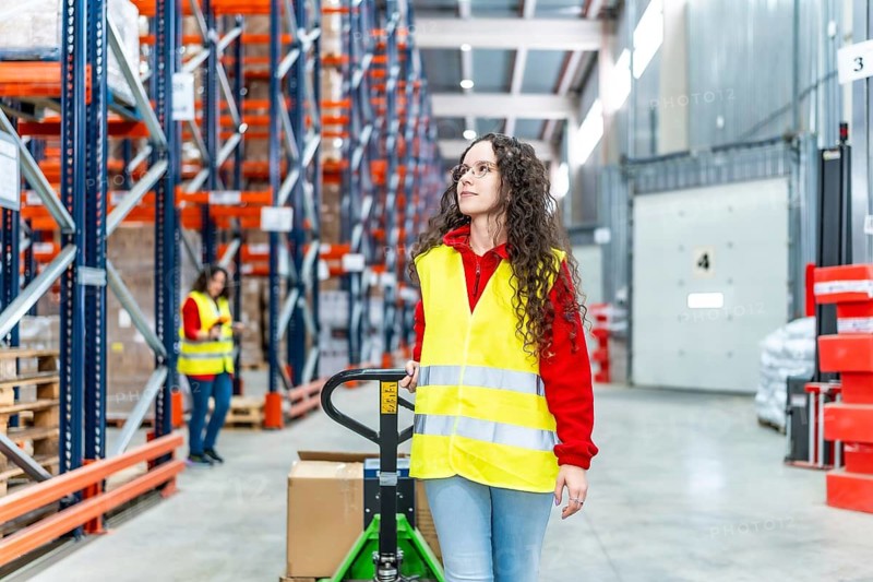 Young caucasian woman worker pushing trolley full of boxes walking along a distribution warehouse corridor