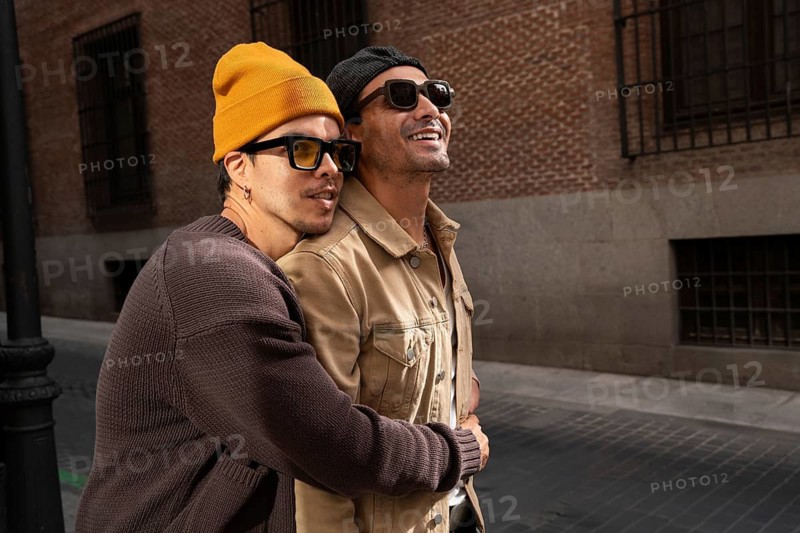 A joyful Colombian couple embraces warmly on a city street, wearing trendy sunglasses and casual clothes, capturing a moment of affection and companionship against a brick wall backdrop