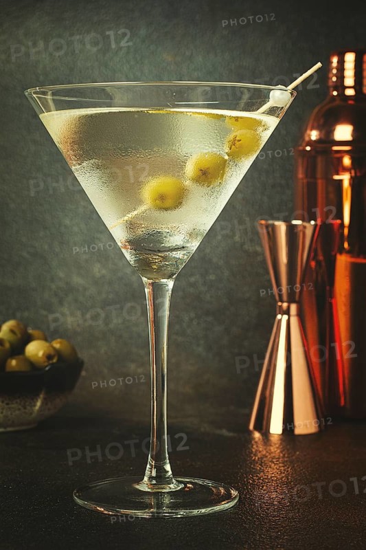 A clear glass holds a martini with olives on a skewer. A cocktail shaker and jigger sit nearby on a dark surface. Green olives rest in a small bowl