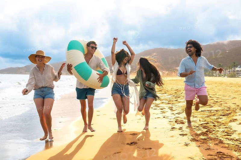 Joyful diverse young friends running barefoot on the sand, laughing and celebrating during a sunny summer vacation on the beach with an inflatable ring