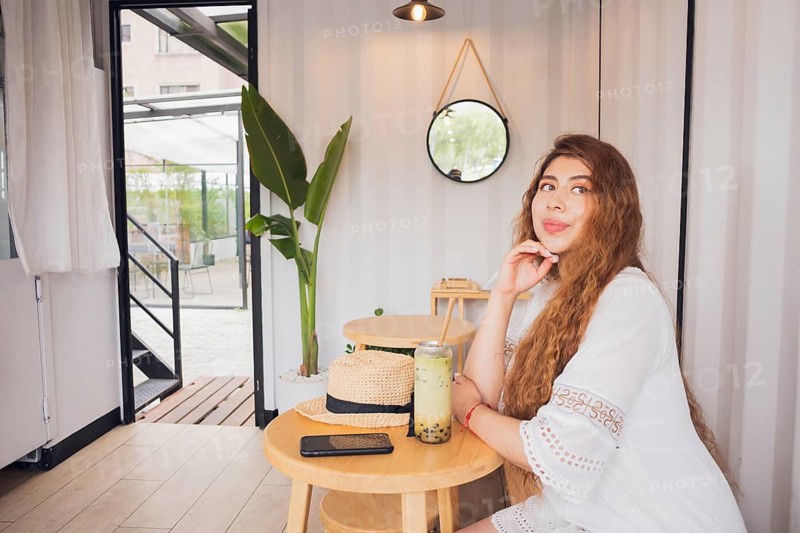 Young American Latin woman enjoying a relaxing lifestyle moment in a modern cafe. She sits at a wooden table with a refreshing drink, wearing a stylish white dress