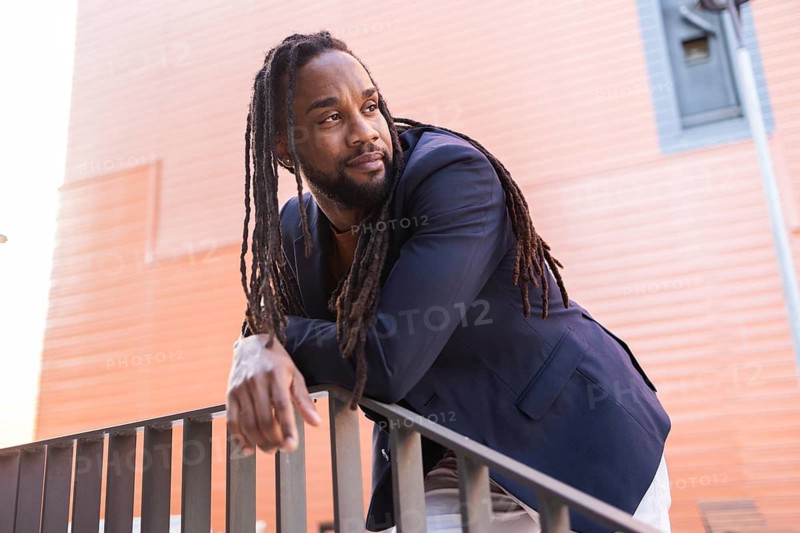 A cuban business man with dreadlocks, wearing a navy blazer, leans thoughtfully on a railing against a modern urban backdrop, conveying a blend of style and contemplation
