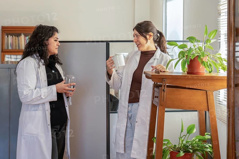 Two young female scientists in white lab coats chatting during a break, one holding a mug and the other a glass of water, standing near a wooden table with potted plants