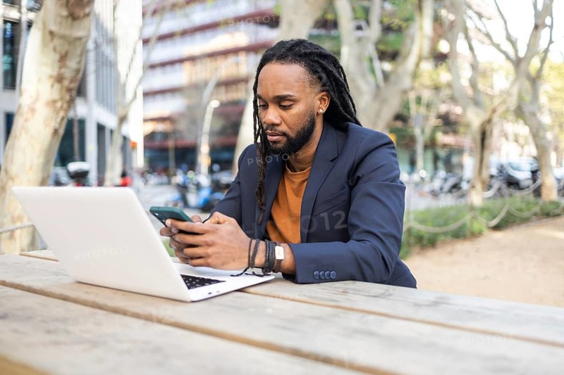 A cuban businessman in a blazer uses a smartphone and laptop while sitting on a wooden bench outside, surrounded by nature and urban elements. Represents remote work and connectivity