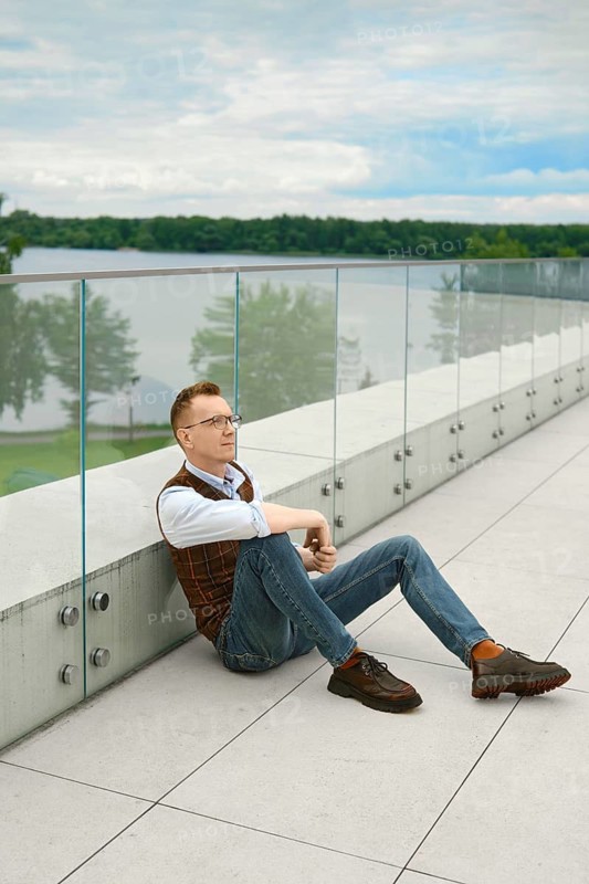 Middle-aged man sits on terrace overlooking lake. He leans back against glass barrier. Calm day with clouds in sky. Surrounding area has trees and green grass. Time appears to be afternoon