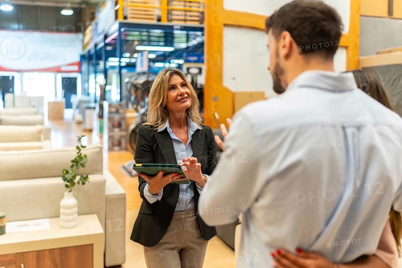 Saleswoman in a suit holding a digital tablet is discussing options with a couple, helping them selecting furniture for their home in a modern store environment