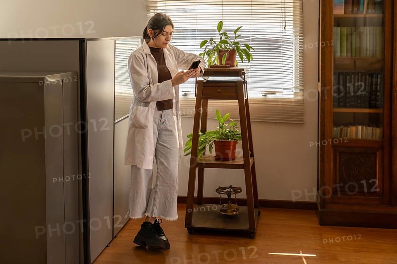 Young female scientist in white lab coat standing near a wooden plant stand with potted plants, looking at her smartphone in a warm sunlit office with filing cabinets and bookcase