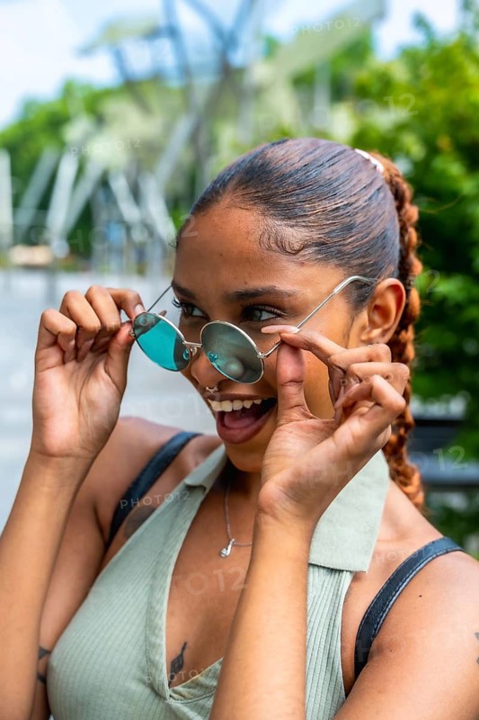 Biracial young woman lowering round sunglasses, looking playfully with a wide smile, expressing happiness and joy while enjoying a vibrant summer day in an urban park