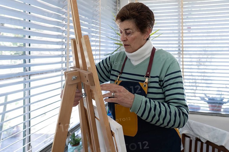 A senior female artist in a denim apron leans over her wooden easel to tighten a screw and secure a canvas in her bright home studio with venetian blinds and houseplants