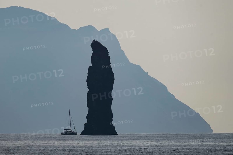 Silhouette of a towering rock needle, behind it the island of Alicudi, a sailboat in the calm sea near Filicudi, Aeolian or Lipari Islands, southern Italy, Italy