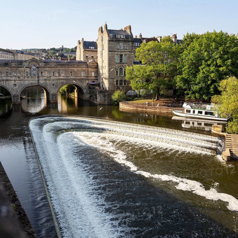 River with small waterfall and bridge, surrounded by old buildings, Bath