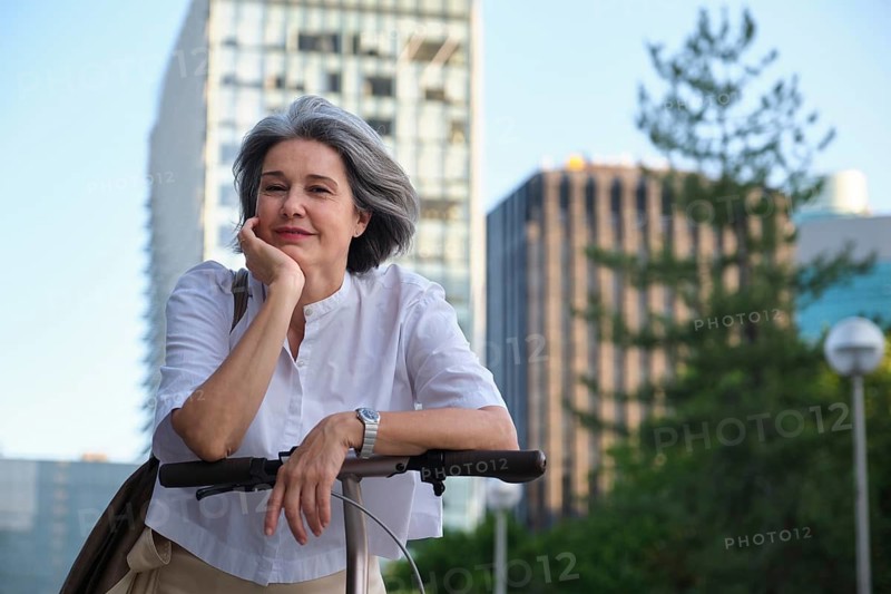 A mature woman with gray hair and a content expression enjoys a leisurely moment on her scooter in an urban setting, with skyscrapers and trees in the background