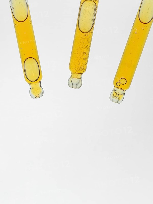 Three glass pipettes for cosmetics filled with amber liquid are suspended against a clear white background. The transparent tubes showcase the fluid and bubbles inside