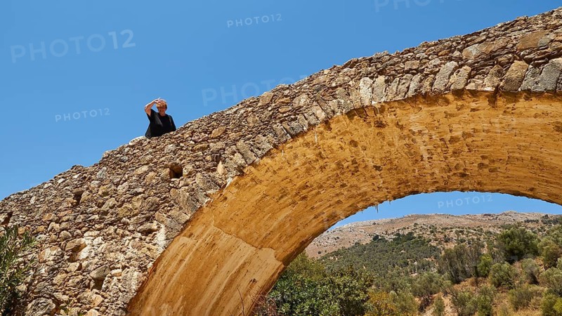 Person on a historic stone bridge looking into the distance surrounded by nature and clear sky, Amari Basin, Amari, Central Crete, Greek Islands, Greece