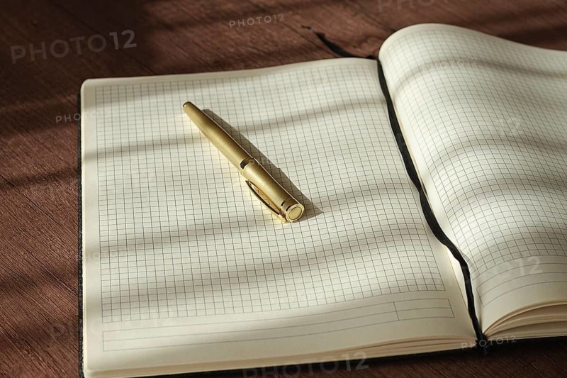 A notebook with blank grid pages is open on a wooden table. A pen rests on the notebook as sunlight creates shadows on the surface. It looks like a space for writing ideas