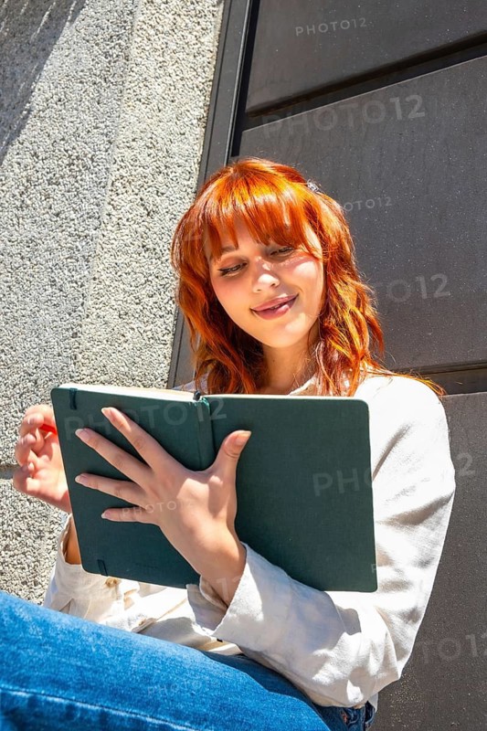 Redhead young woman smiling while writing or sketching in a green notebook, finding inspiration and expressing creativity outdoors during a bright summer day