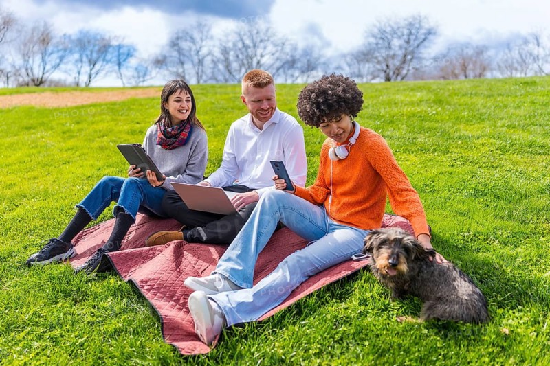 Three students are using electronic devices while relaxing on a blanket in a park with their dog