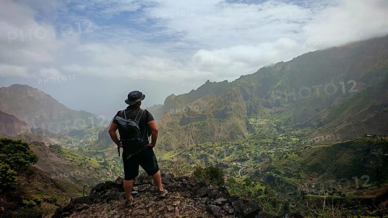 Male person with backpack stands on a rock and looks into the valley surrounded by green mountains and enjoys the view, hiking from Cova Crater through Paul Valley, Ribeira do Paul to Vila das Pombas, Santo Antao, Cabo Verde, Cape Verde, Cape Verde