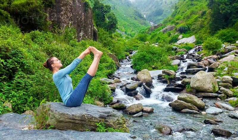 Yoga exercise outdoors, panorama of woman doing Ashtanga Vinyasa Yoga balance asana Ubhaya padangusthasana Big Double Toe Yoga Pose at tropical waterfall in Himalayas in India. Panoramic image