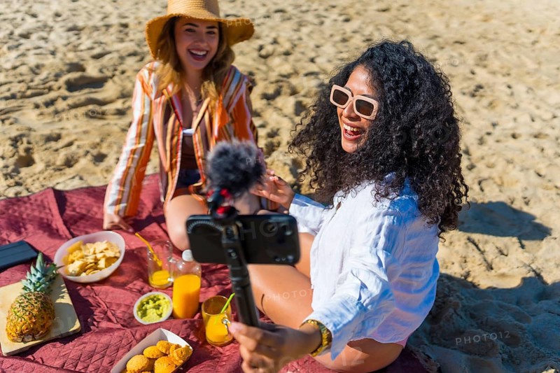 Two cheerful young women, one latina with curly hair holding a smartphone on a tripod with a microphone, creating a vlog or livestream while enjoying a summer beach picnic
