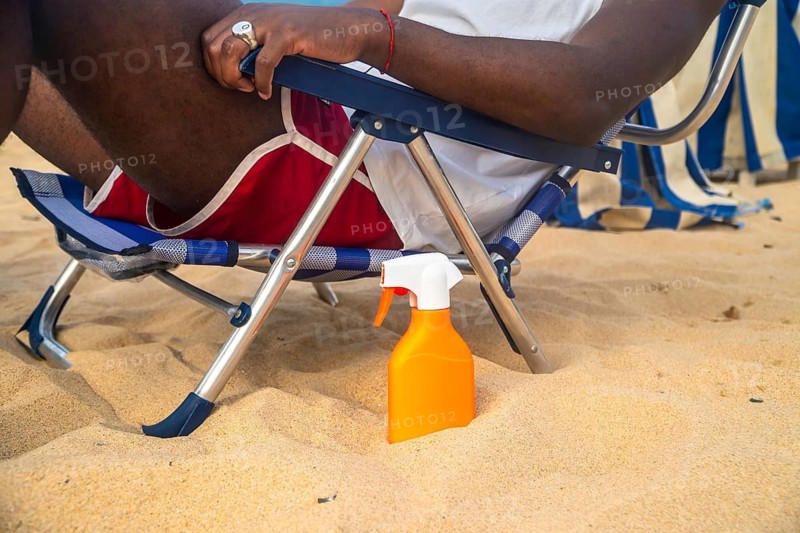 Person enjoying a sunny day at the beach, sitting on a chair with an orange sunscreen spray bottle protecting skin from sunlight, experiencing leisure during a holiday trip