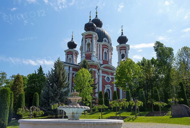 Orthodox church with red and white accents surrounded by trees against a blue sky with a fountain in the foreground, Cathedral of the Nativity of the Virgin Mary, Curchi monastery, Orhei district, Republic of Moldova, Moldova