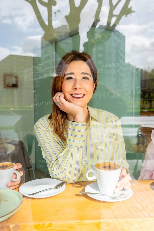 Young woman leaning on hand and smiling, looking out from a cafe window with reflections while enjoying a hot coffee, representing leisure and a cozy lifestyle