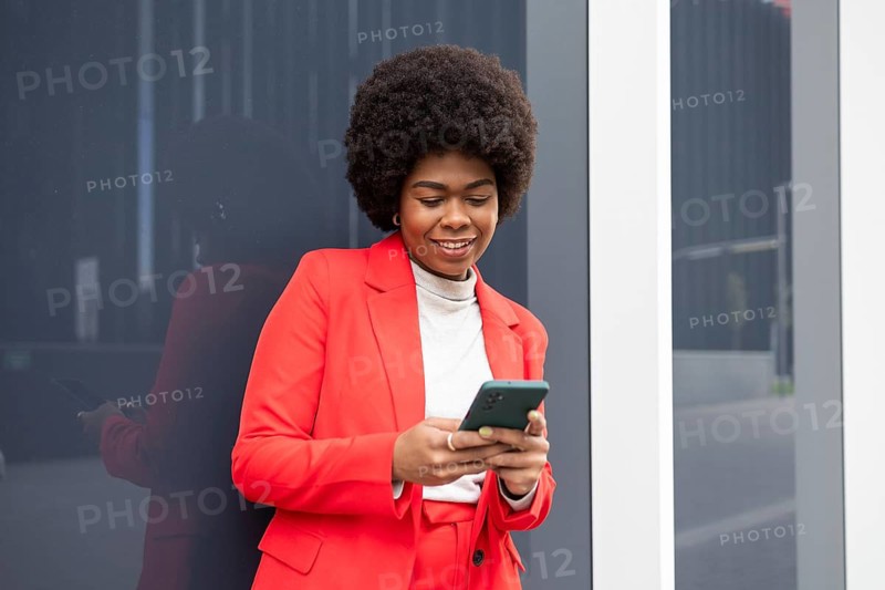 A confident woman in a bright red suit using a smartphone while standing outside by a reflective surface, showcasing modern urban lifestyle and fashion